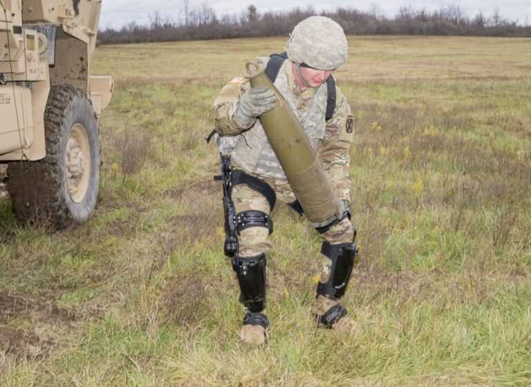 A soldier in camouflage uniform and protective gear is lifting a large artillery shell while wearing a lower-body exoskeleton in an open grassy field. The exoskeleton, consisting of mechanical braces on his legs, appears to assist with mobility and heavy lifting, reducing strain on his body. A military vehicle with rugged tires is partially visible in the background, suggesting that the scene is part of a field operation or a demonstration of exoskeleton-assisted soldier performance.