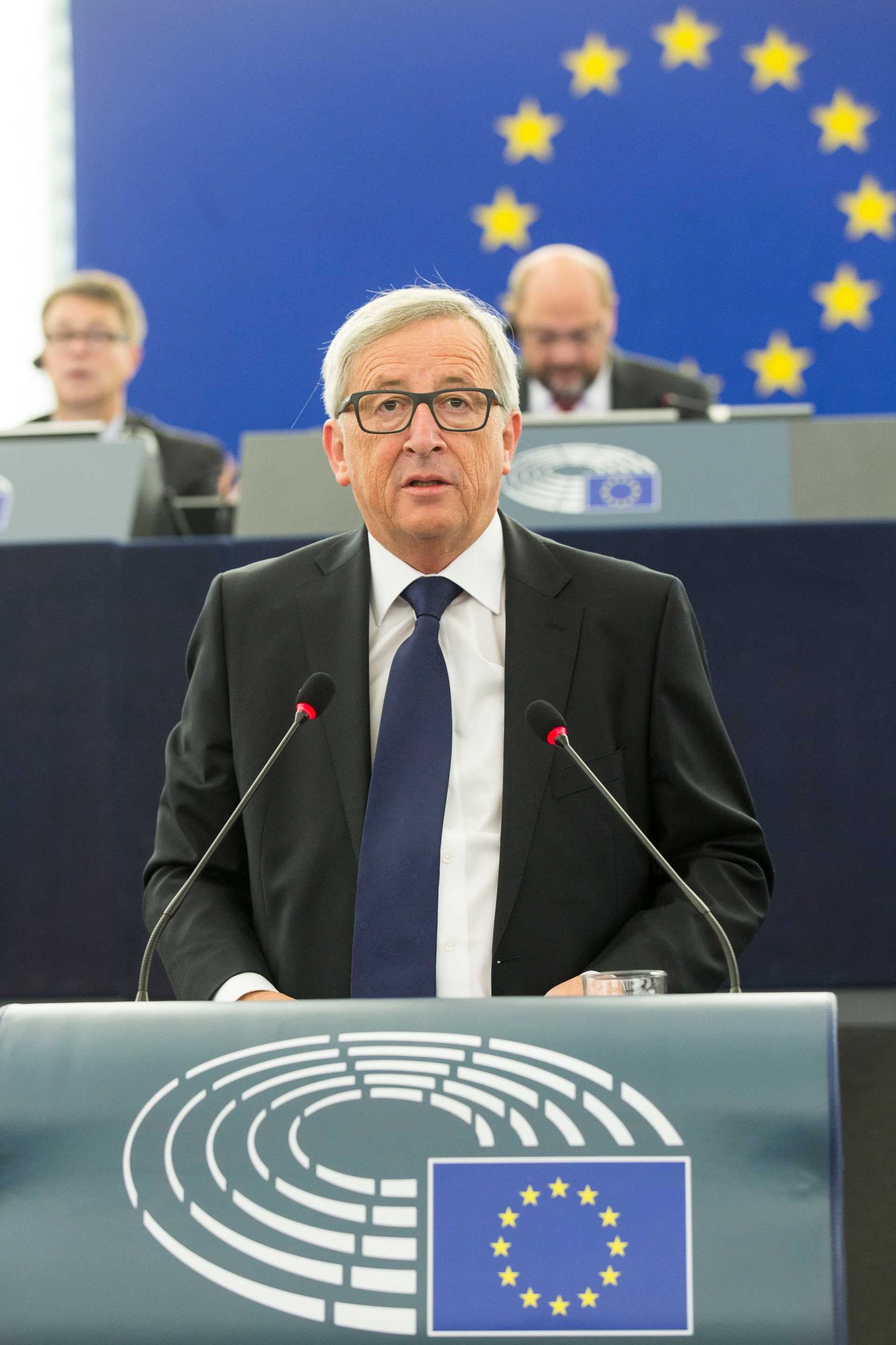 Jean-Claude Juncker in parliament behind a speaker's desk. His facial expression is serious, so is his clothing with suit and tie. In the background is a European Union flag.
