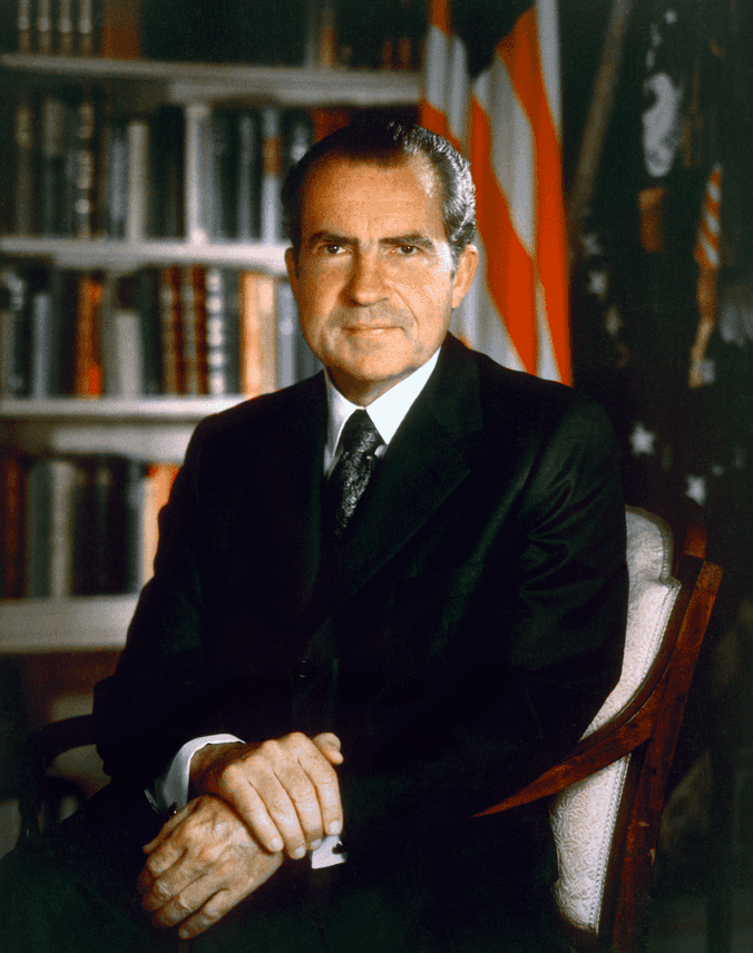 Portrait of the US President Richard Nixon in color. He is wearing a suit and is sitting in front of a bookshelf and an US flag.