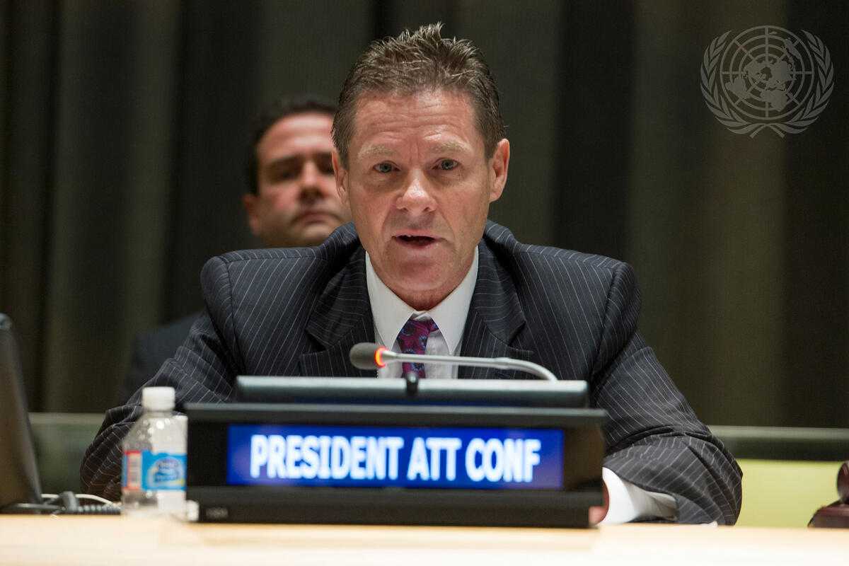 The image shows a man in a dark pinstripe suit, white shirt, and patterned tie speaking into a microphone at a formal conference. He has short, neatly styled hair and a serious expression. In front of him is a nameplate with the illuminated text 'PRESIDENT ATT CONF', 