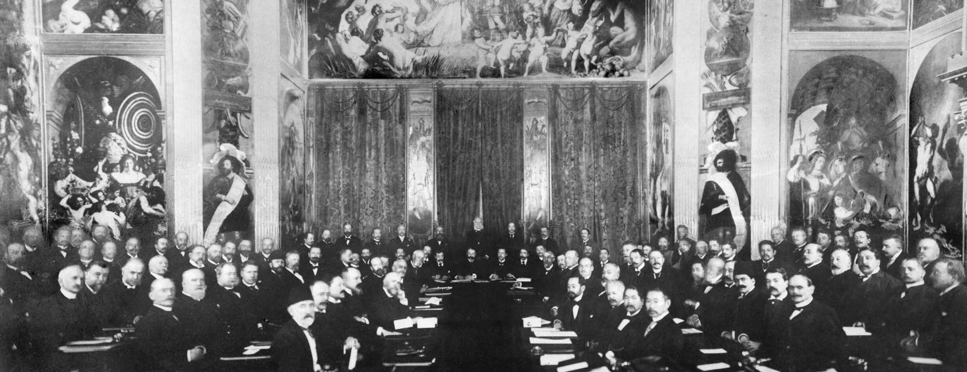 Historic photo of a conference. The room is decorated with murals. Men in dark suits sit in rows and look towards the camera.