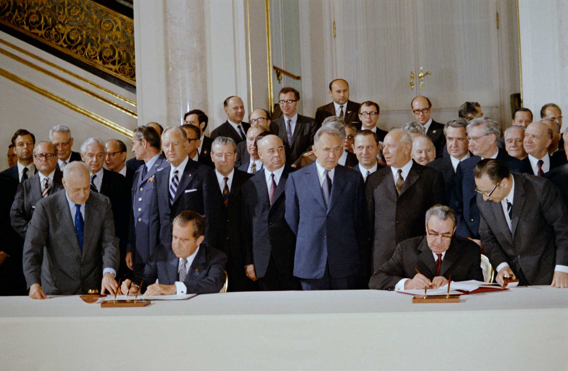 President Nixon (USA) and General Secretary Brezhnev (USSR) sitting at a table signing documents, with more than 30 white men standing in the back watching.