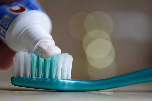 A close-up photograph of a turquoise toothbrush with white and green bristles. A dollop of white toothpaste with small red specks is being squeezed onto the bristles from a partially visible toothpaste tube. The background is blurred, with soft circular light reflections creating a bokeh effect.