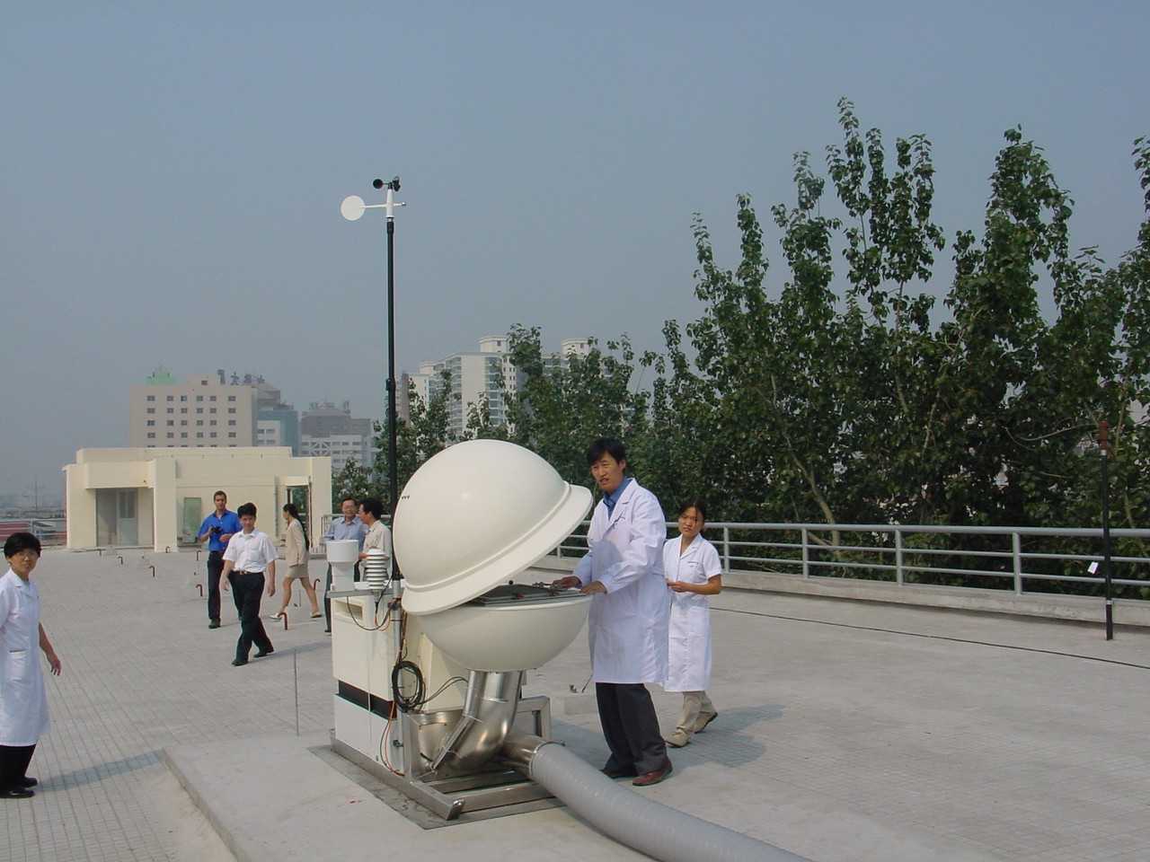 The image shows a radionuclide monitoring station in Beijing, part of the CTBTO’s International Monitoring System, which detects radioactive particles from nuclear explosions. In the foreground, a man in a white lab coat stands next to a large white spherical device with its top half open, revealing monitoring equipment inside. Several metal tubes and a thick hose are connected to the base. Other people in lab coats and casual clothing are walking or observing the scene on the rooftop of a building. Trees and a city skyline with tall buildings are visible in the background under a hazy sky. The atmosphere appears calm and professional