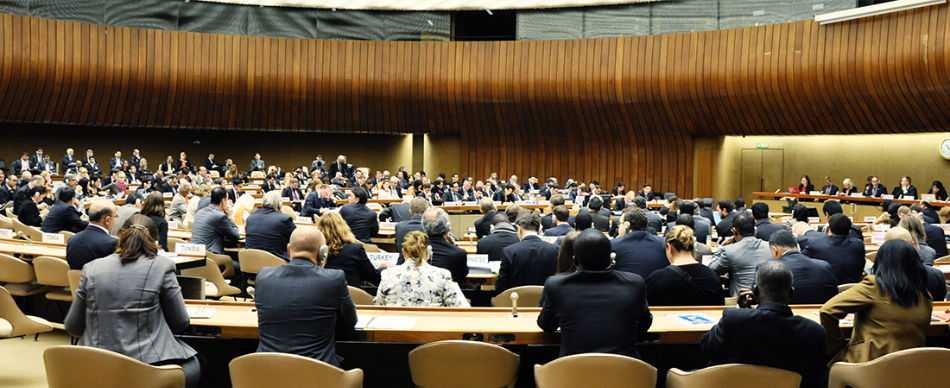 Backs of people sitting in rows of seats, attending a conference in a huge conference room.