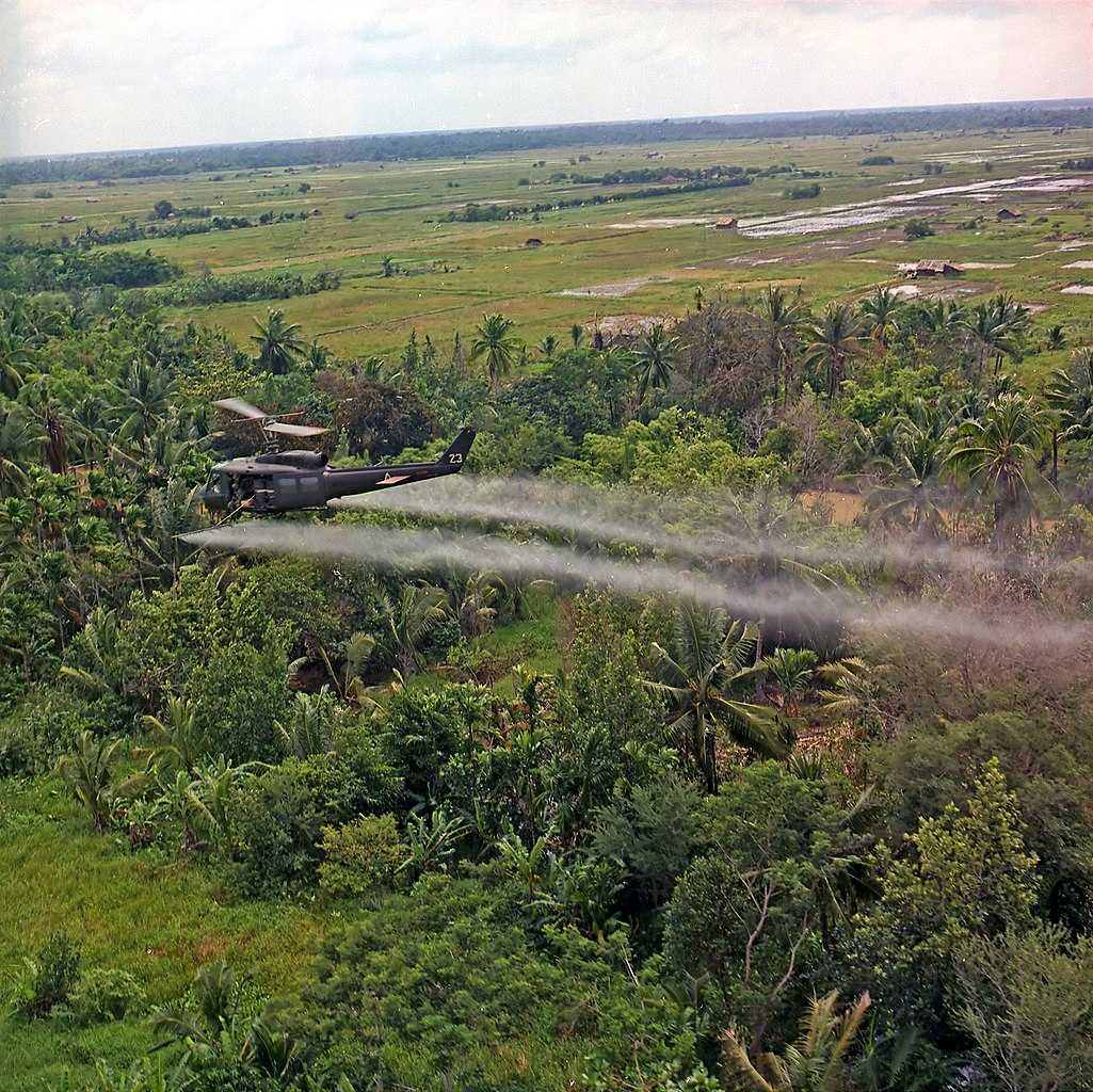 Photograph depicting an UH-1D helicopter in flight, spraying a defoliation agent on agricultural land