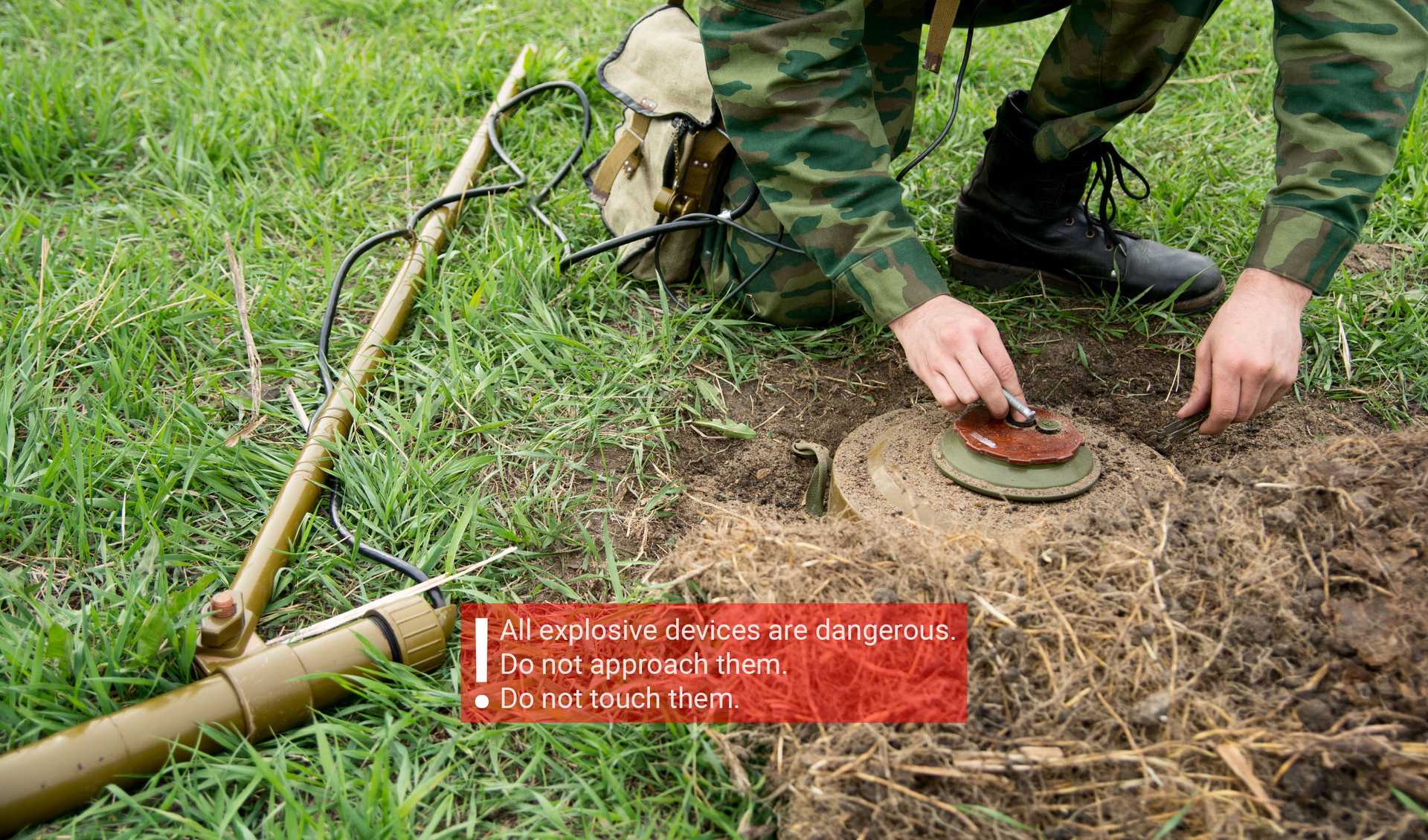 A soldier is carefully handling a landmine partially buried in the ground, using a metal detector and tools. The surrounding area is grassy, and a warning sign in the image reads: All explosive devices are dangerous. Do not approach them. Do not touch them.