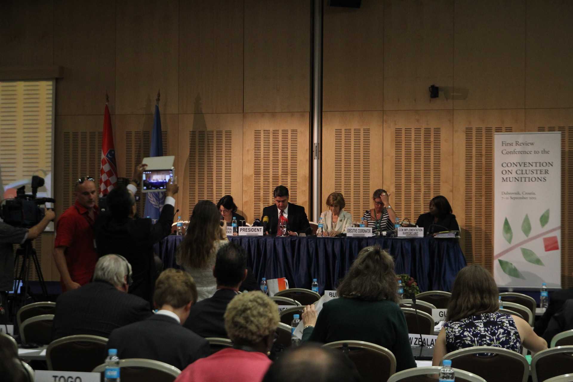 A formal conference setting with a panel of six individuals seated at a long table on a stage. A man in a dark suit and red tie sits in the center with a nameplate reading 'PRESIDENT,' appearing to lead the discussion.