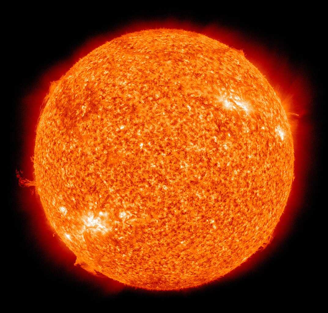 Image of the Sun, glowing intensely in shades of orange and red. The surface appears textured with swirling plasma patterns and bright flares. Prominences extend from the edges, and a faint red halo surrounds the Sun. The image captures the dynamic, fiery nature of our star in vivid detail.