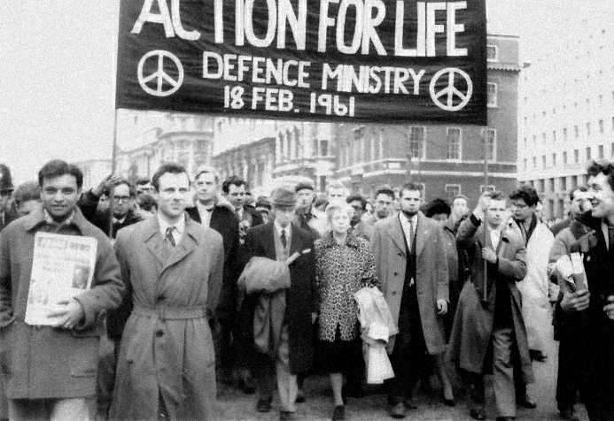 Historic photo of people in coats, suits and dresses marching. They hold a banner that shows two peace signs and reads: Action for Life. Defence Ministry, eighteenth February 1961.