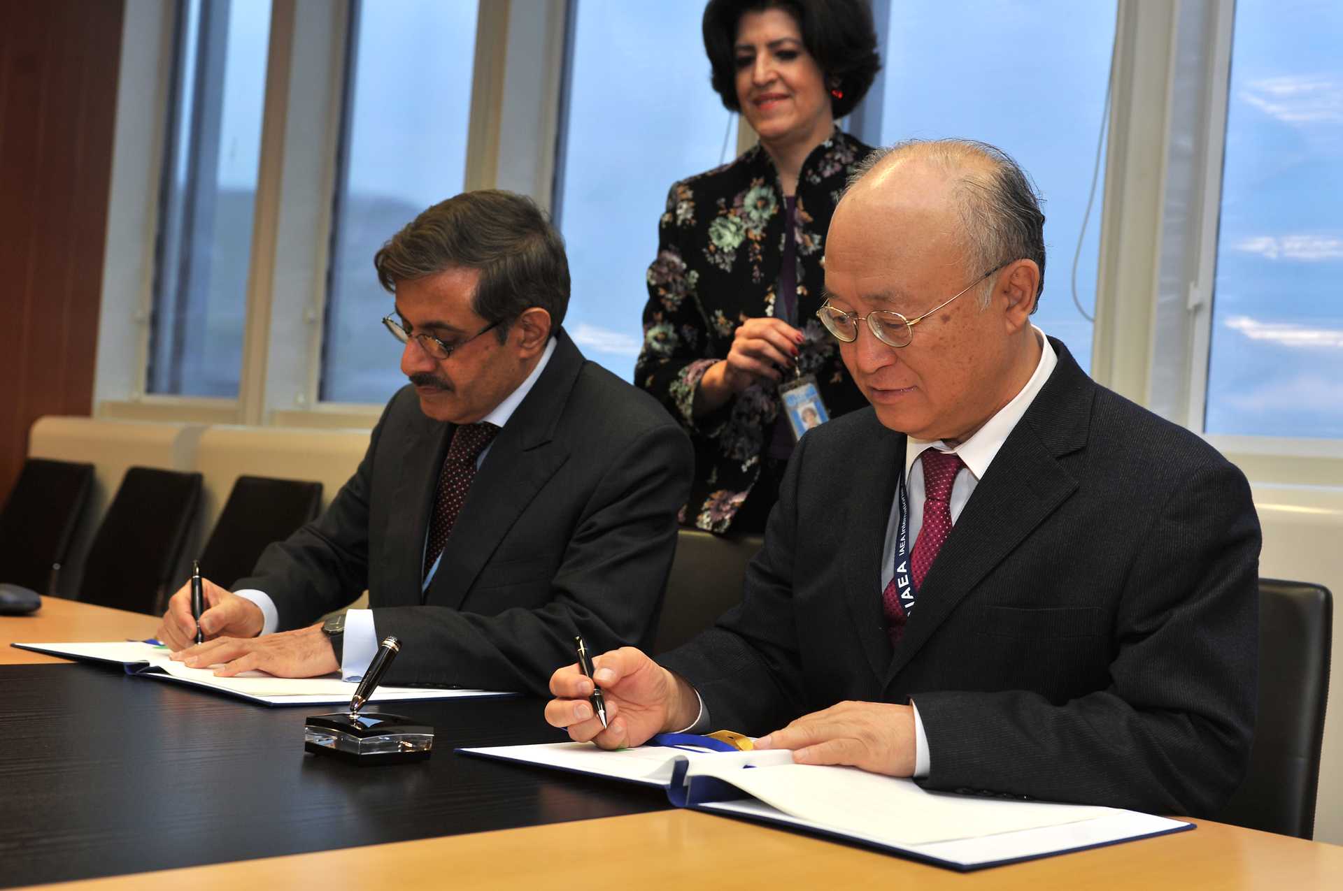 Two men in suits sit at a conference table and sign documents. A bystander watches.