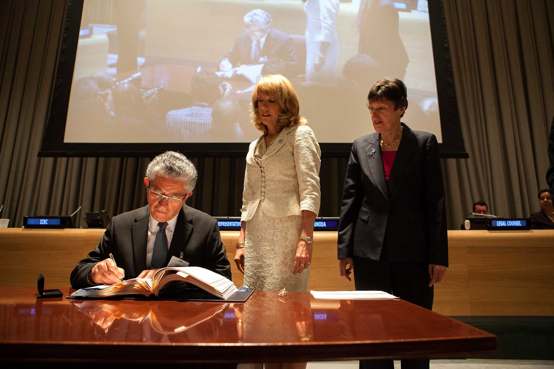 A man in a dark suit and glasses is seated at a table, signing a large official document with a pen. Two women, one in a white outfit and the other in a dark suit, stand beside him, observing the signing. The setting appears to be a formal conference room or an official institution, with a large screen in the background displaying a live feed of the event. The atmosphere suggests a significant diplomatic or governmental signing ceremony