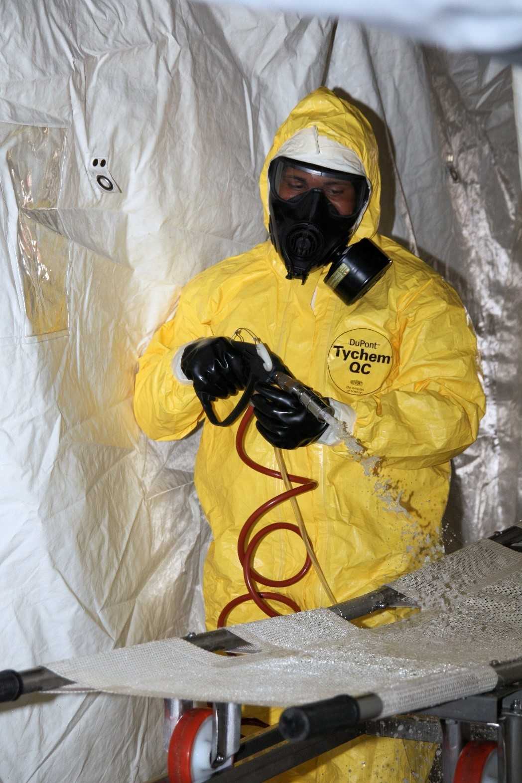 Scene of a decontamination process, showing a person in a yellow hazmat suit and black gloves using a hose to clean a surface.