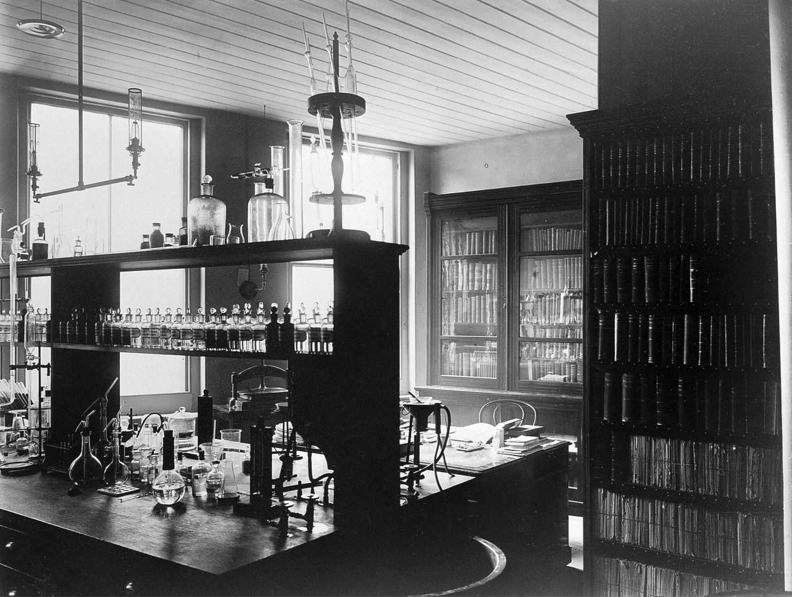 Historic photograph of the interior of a pharmaceutical or chemical labaratory with a bench in the centre and books annd papers on shelves around the walls.