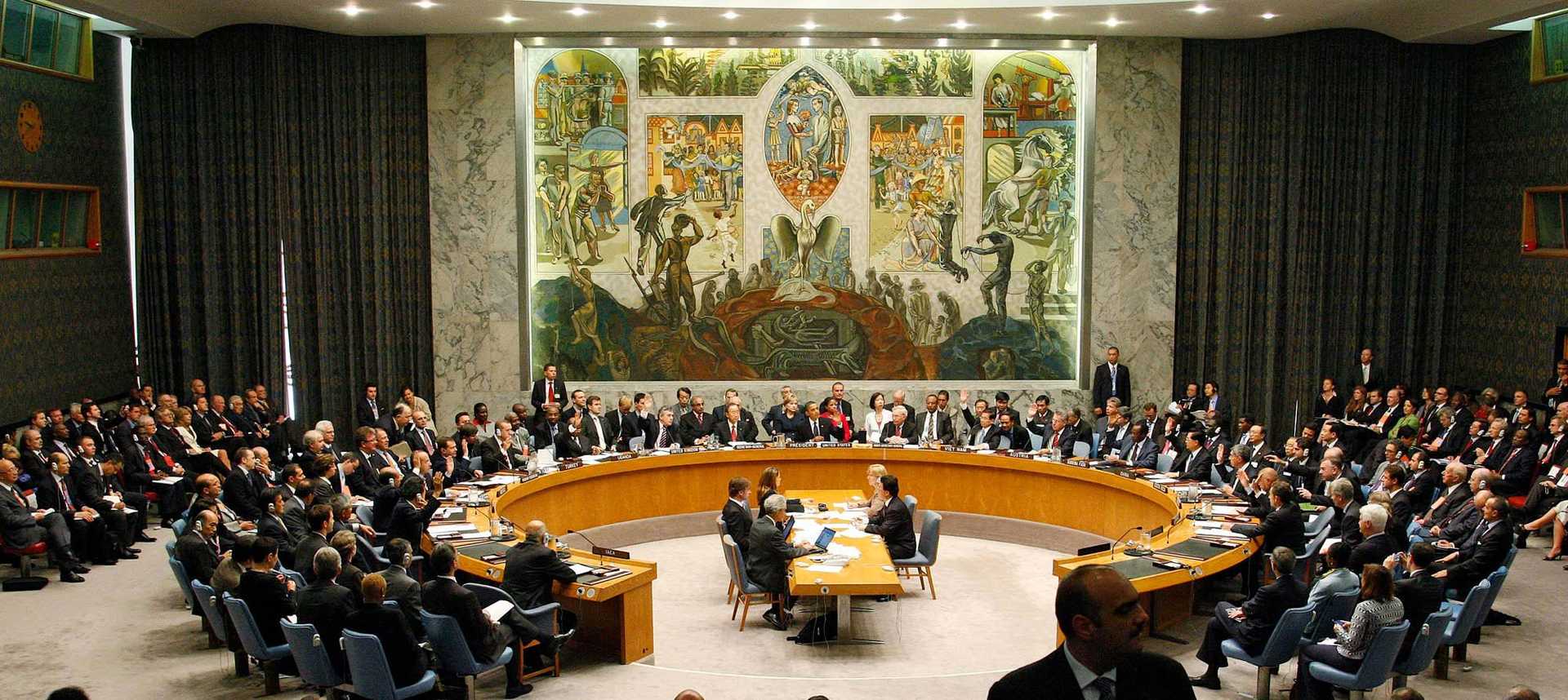 Photo of a meeting in a highly decorated conference room. Tables arranged in a circle surround a small rectangular table. More rows of chairs filled with people around the tables.