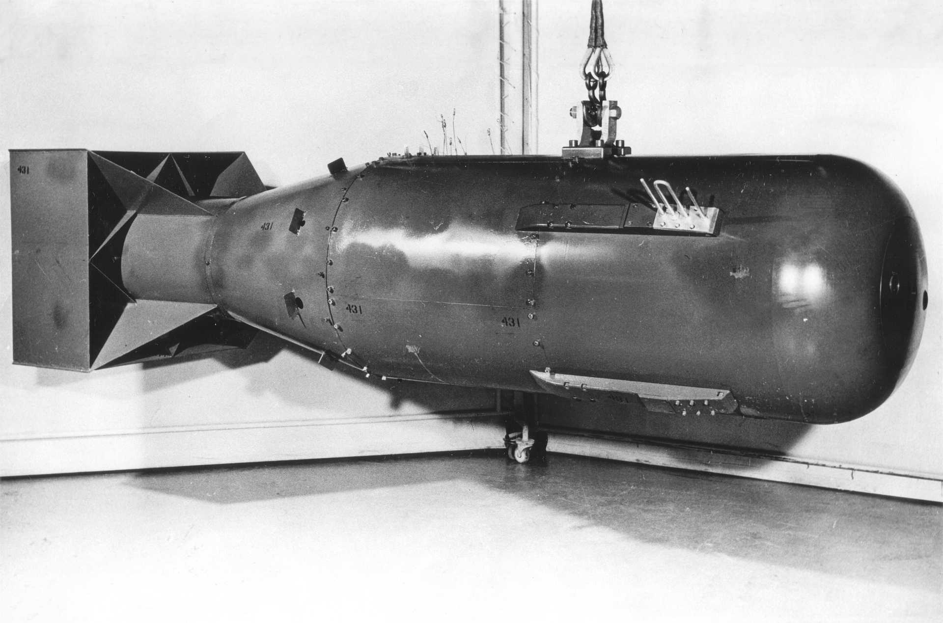 Black-and-white photo of a large, cylindrical bomb suspended by a hook in a room. The device has a rounded nose, stabilizing fins at the back, and visible panels and fasteners along the body. This is a historical image of the „Little Boy“ bomb, shown in a controlled indoor setting. 
