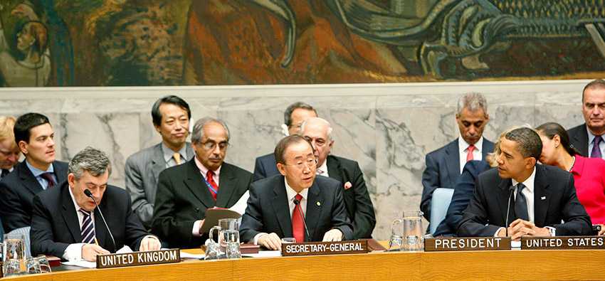 Photo of people at a meeting, they sit side by side on a table and have name tags: United Kingdom, Secretary-General, President United States.