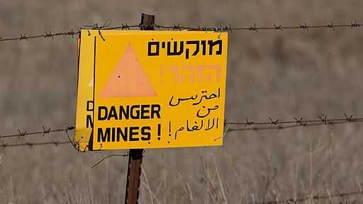 The image shows a yellow warning sign attached to a barbed wire fence in a dry, open landscape. The sign warns of landmines in Hebrew, English, and Arabic. The English text reads 'DANGER MINES!'. The Hebrew text at the top says 'מוקשים' (Mokshim), meaning 'Mines', and 'הזהר!' (Hizaher!), meaning 'Beware!'. The Arabic text reads 'احترس من الألغام!' (Ihtaris min al-algham!), which translates to 'Beware of the mines!'. A red triangle symbol reinforces the warning. The background consists of dry grass or barren land, suggesting a hazardous, possibly former conflict zone or military area. The sign serves as a critical alert to prevent people from entering the dangerous mined area.
