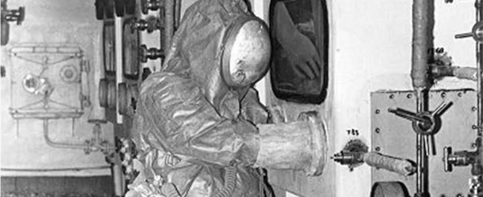 Historic photo of a person all covered in a hazard suit with unspecific machinery around.