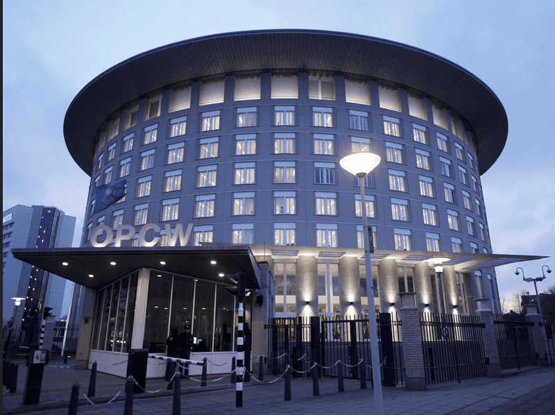 OPCW headquarters building in The Hague - a modern cylindrical structure with illuminated windows at dusk, featuring a glass entrance canopy with 'OPCW' signage and security barriers.