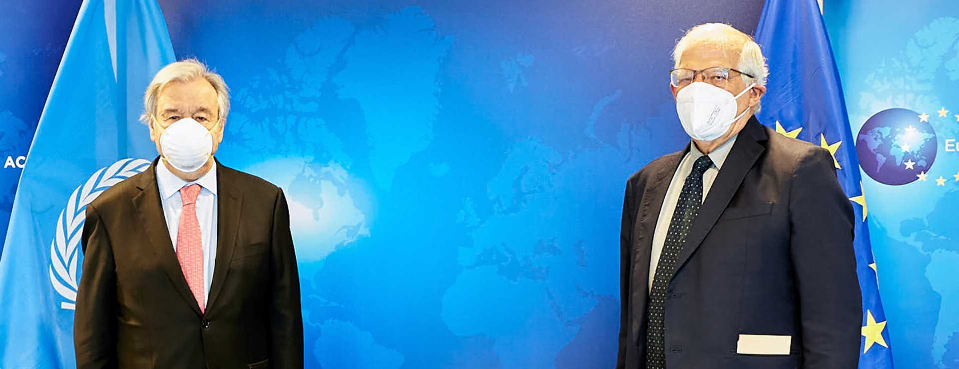 Two men with grey hair and dark suits stand in front of a blue background featuring the EU and the UN flag. They wear face masks and keep distance to each other.