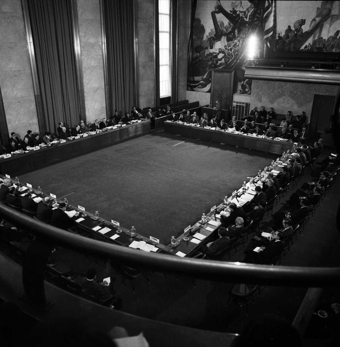 Black and white picture of a huge square of tables from above. On the tables lie documents. People sit on them, attending a conference.