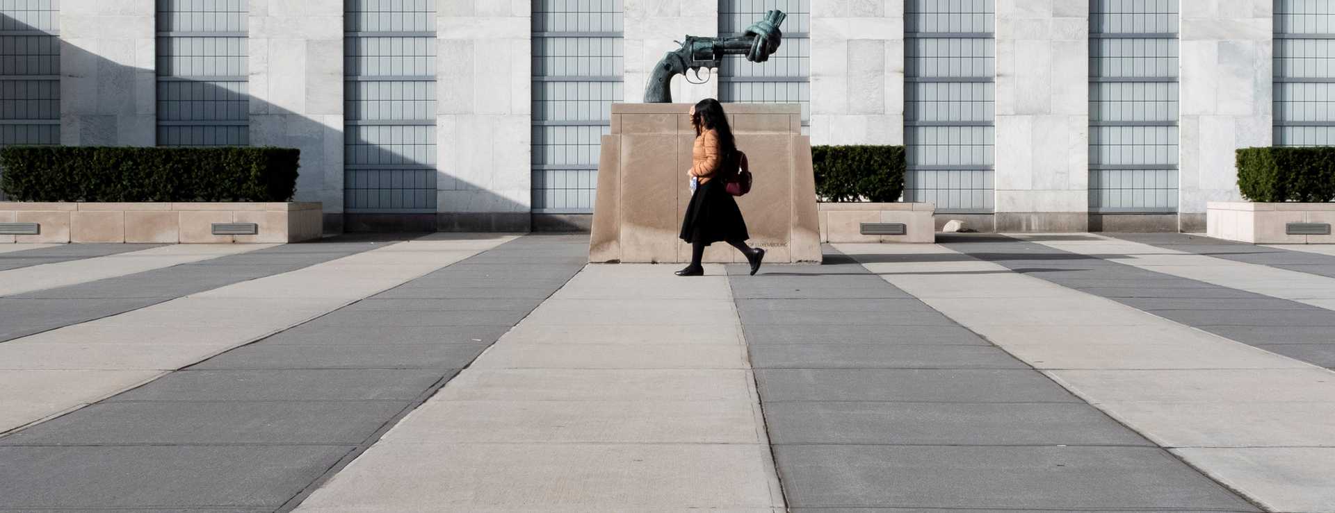 Photo of a person who walks by the statue of a knotted gun displayed in front of a bulding's facade.