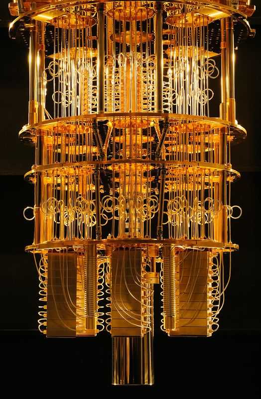 The image showcases a quantum computer's dilution refrigerator, a critical component used to cool quantum processors to extremely low temperatures—close to absolute zero. The structure is highly intricate and composed of multiple circular and cylindrical layers interconnected by thin golden wires and vertical rods. These layers are arranged in a symmetrical fashion, giving the apparatus a futuristic and visually striking appearance. The golden color of the metal components stands out against the dark background, enhancing the overall aesthetic appeal. This cooling system is essential for maintaining the stability of qubits, the fundamental units of quantum computation, by reducing thermal noise and preserving quantum coherence.