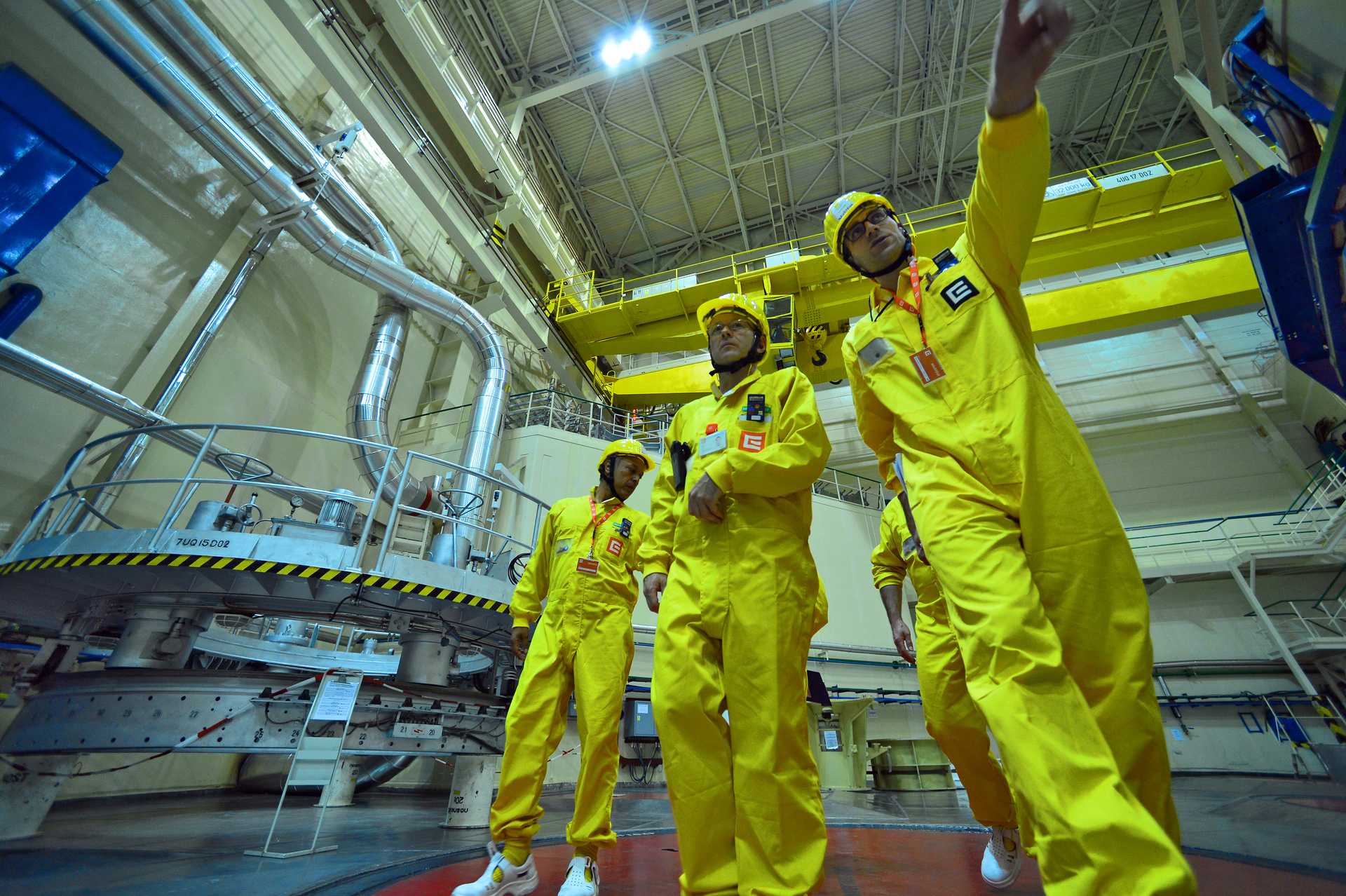 Four individuals wearing bright yellow protective suits and helmets walk through an industrial facility. One person in the foreground is pointing forward. The background features large machinery, pipes, and metal structures inside a nuclear power plant. The image was taken during a 2015 Safeguards Comprehensive Training Exercise at Dukovany Nuclear Power Plant in the Czech Republic 