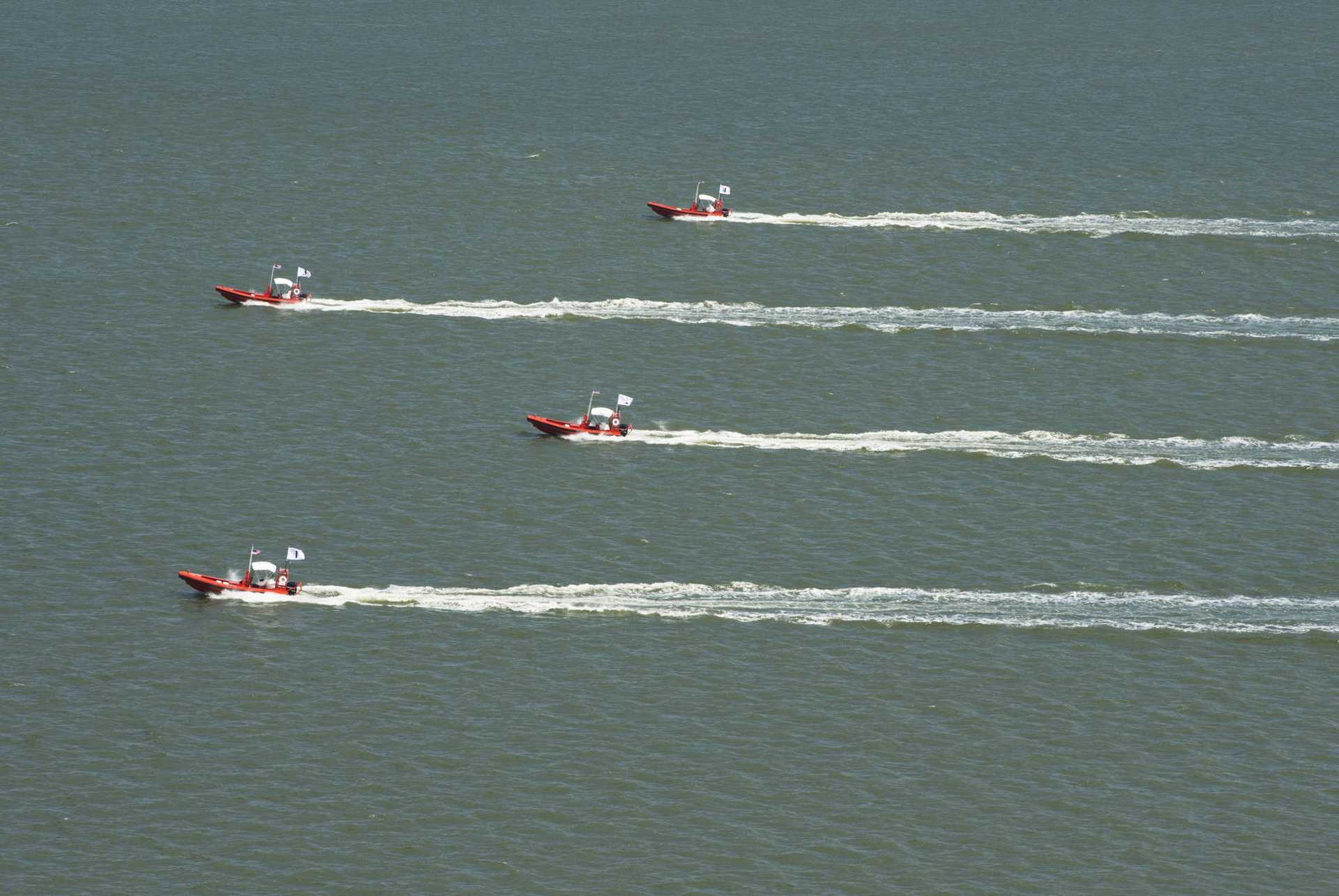 Aerial view of four boats travelling in a slightly staggered formation next to each other. The previous course of the boats can be clearly seen through the churned-up water behind them and runs parallel.