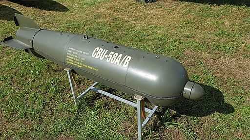 The image shows a CBU-58A/B cluster bomb, a military ordnance painted in dark green, displayed outdoors on a metal stand on a grassy surface. The bomb is cylindrical with a rounded nose and stabilizing fins at the rear. Yellow markings on the side indicate identification details, including the designation 'CBU-58A/B'. This type of weapon is designed to release multiple smaller submunitions upon deployment. The setting suggests that the bomb is deactivated and displayed as part of a museum or exhibition.