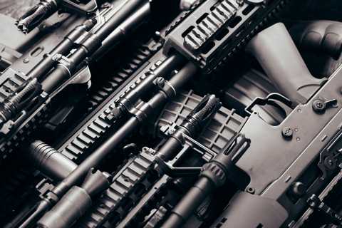 A close-up black and white photograph of multiple modern firearms stacked together, showing detailed views of barrels, rail systems, grips, and various attachments.