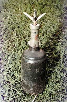 The image shows a POM-1 anti-personnel fragmentation mine standing upright on a grassy surface. The mine consists of a cylindrical metal body with a fuse mechanism on top, which has multiple metallic prongs extending outward. The body appears dark and metallic, showing signs of wear or exposure to the elements. This type of mine is designed to scatter shrapnel upon detonation, posing a significant risk to anyone nearby. The mine is set in a natural outdoor setting, likely indicating it is in a field or conflict-affected area. 