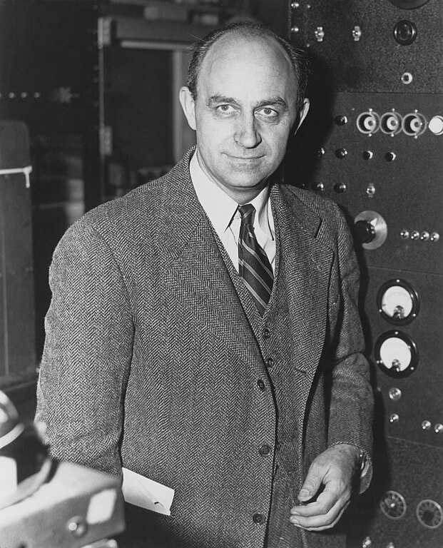 Black and white portrait of Enrico Fermi, a key figure in nuclear science. He stands confidently in a suit and tie, holding a piece of paper, with scientific equipment featuring dials and switches behind him. His expression is focused, reflecting his role in pioneering nuclear reactor development and atomic research.