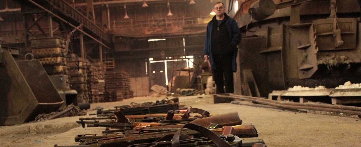 A man stands in a storage hall. Various rifles are stacked up on the floor in front of him.