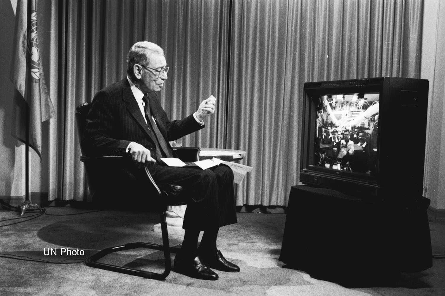 Black and white photo of Secretary-General Boutros Boutros-Ghali sitting in front of a TV with the UN flag in the background, celebrating the 50th anniversary of the UN with the Shuttle-Mir docking mission on November 17, 1995
