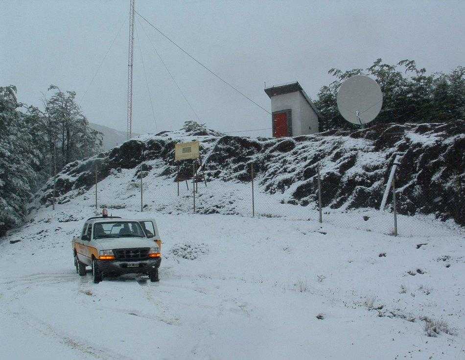 The image shows a remote seismic monitoring station operated as part of the CTBTO’s International Monitoring System, which detects underground nuclear tests. A white pickup truck with orange stripes and an open driver-side door is parked on a snow-covered path in the foreground. Snow blankets the ground, nearby trees, and rocky terrain. A fenced-off area on a small hill contains a white building with a red door, a large satellite dish, and a tall communications antenna. A yellow sign is attached to the fence, but the text is unreadable. The scene appears cold and overcast, suggesting a winter day.