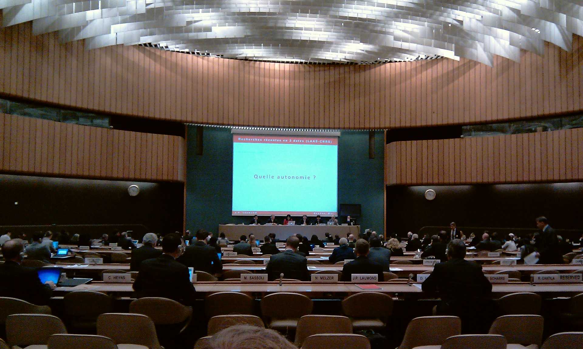 icture taken from far back in a large conference room. You can see many people from behind, looking in the direction of a stage and very large projector screen in the middle of the picture. In front of the screen on the stage you can see a small panel with several people who cannot be individually identified.
