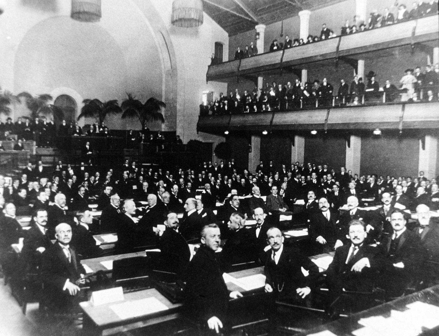 Historic photo of the League of Nations. In a huge hall, many men in dark suits attend a conference. They are sitting in rows as well as standing on balconies and facing the viewer.