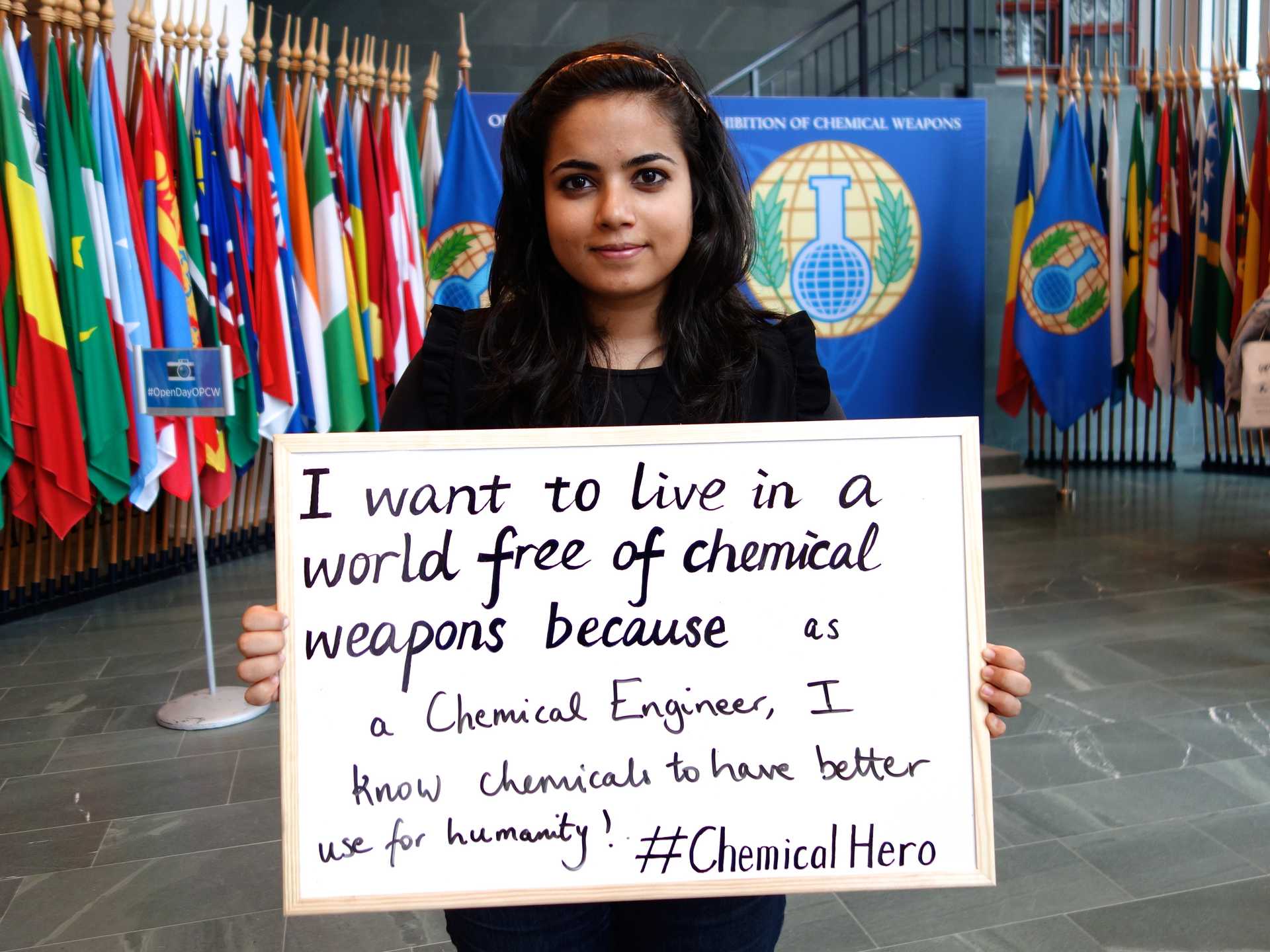 Picture of a young women standing in front of several flags and holding a sign that says 'I want to live in a world free of chemical weapons because as a Chemical Engineer, I know chemicals to have better use for humanity. #ChemicalHero'. The hashtag identifies the action as part of a campaign.