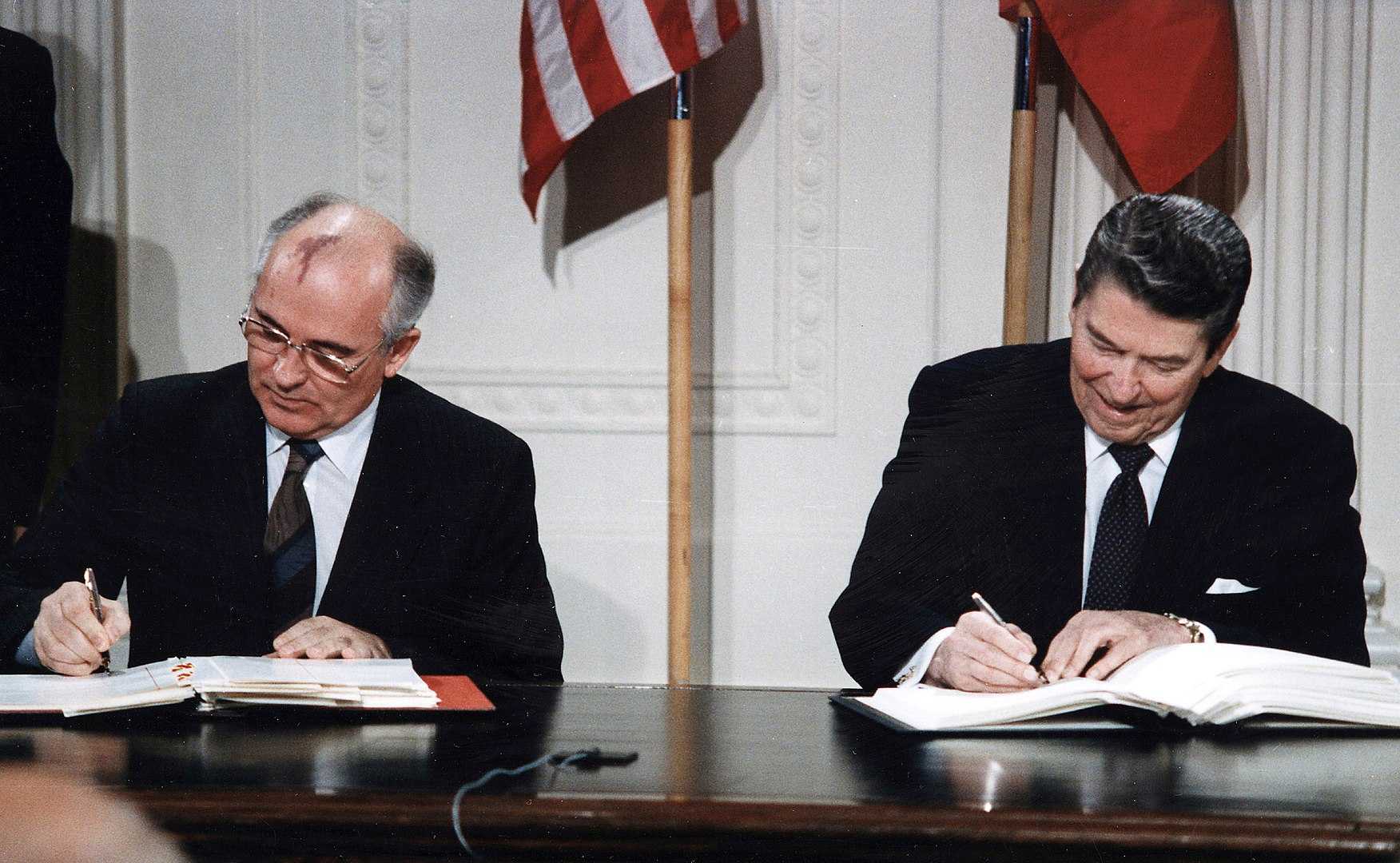 Secretary General Gorbachev and President Reagan sitting on a wooden desk with the American and Soviet flag in the back. Each of them has a folder in front of them in which they sign something. 