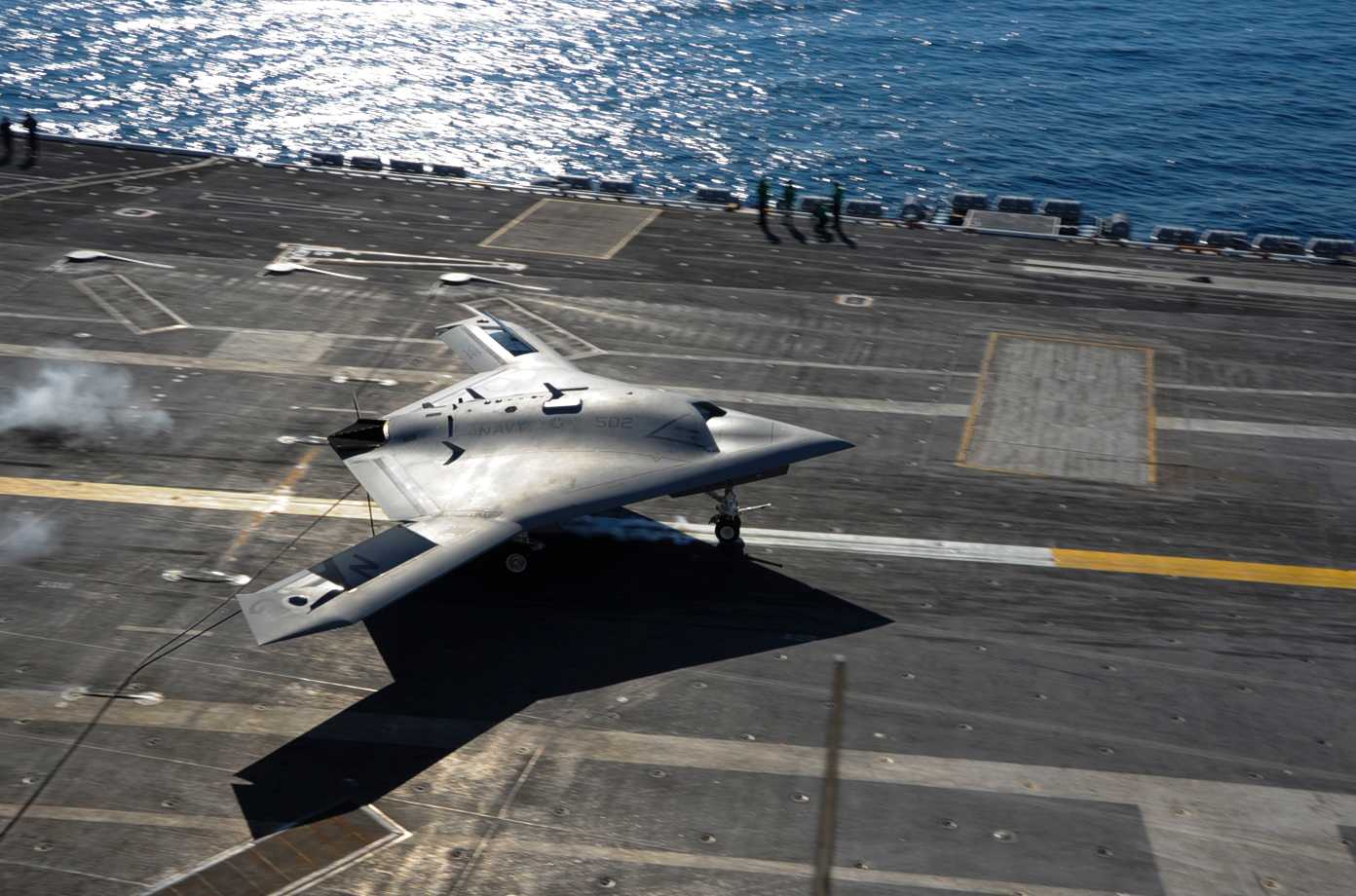 A photo taken from an angle above shows a strangely shaped aircraft without a cockpit on an aircraft carrier. You can see water in the background. People in the background appear small compared to the aircraft, but the size of the aircraft is difficult to estimate.