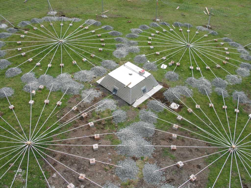 Aerial view of a circular infrasound sensor array with multiple white rods radiating from several central hubs, connected to sensors anchored by small blocks and surrounded by patches of gravel, all arranged around a small, bunker-like building on a grassy field enclosed by a temporary fence.