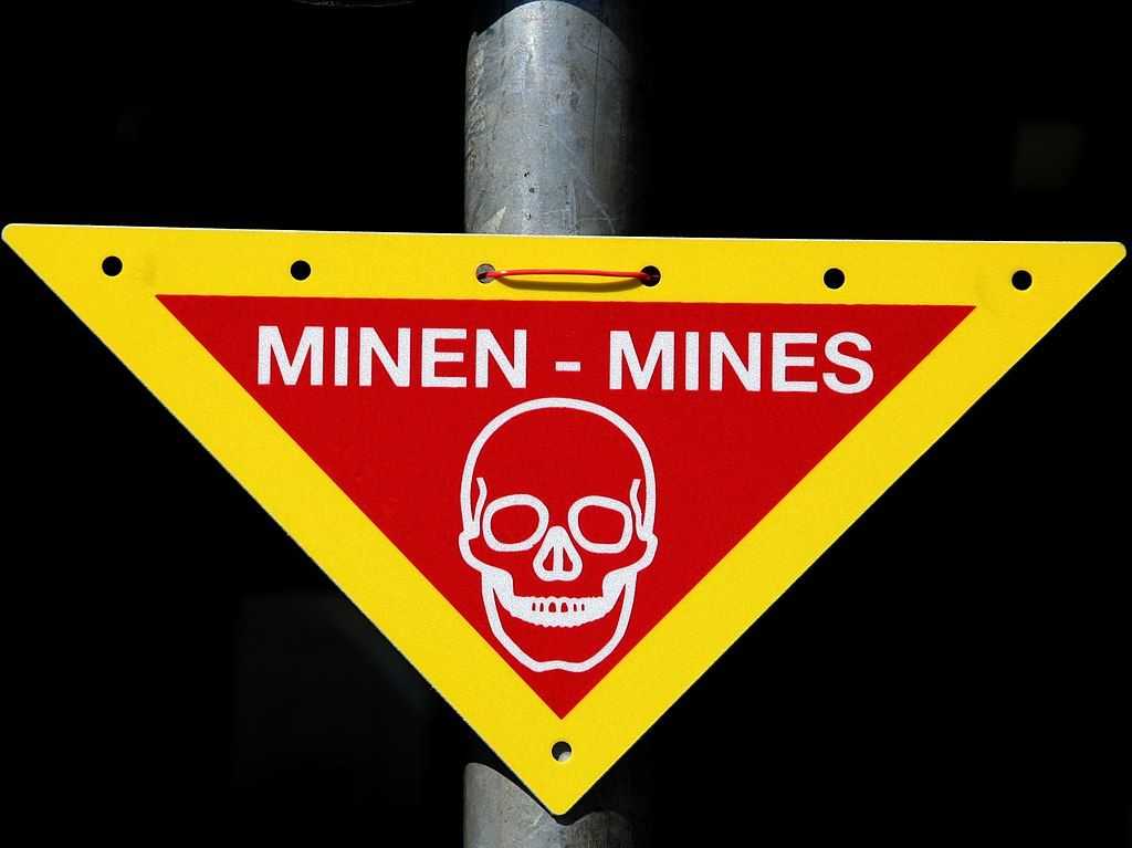 A triangular warning sign with a yellow border and a red center is attached to a metal pole. The sign prominently displays the word 'MINEN - MINES' in white capital letters, warning of landmines. Below the text, there is a white skull symbol, reinforcing the danger. The sign is well-maintained and clearly visible against a dark background. This type of sign is typically used in military or former conflict zones to alert people to the presence of landmines and prevent accidental entry into hazardous areas. 