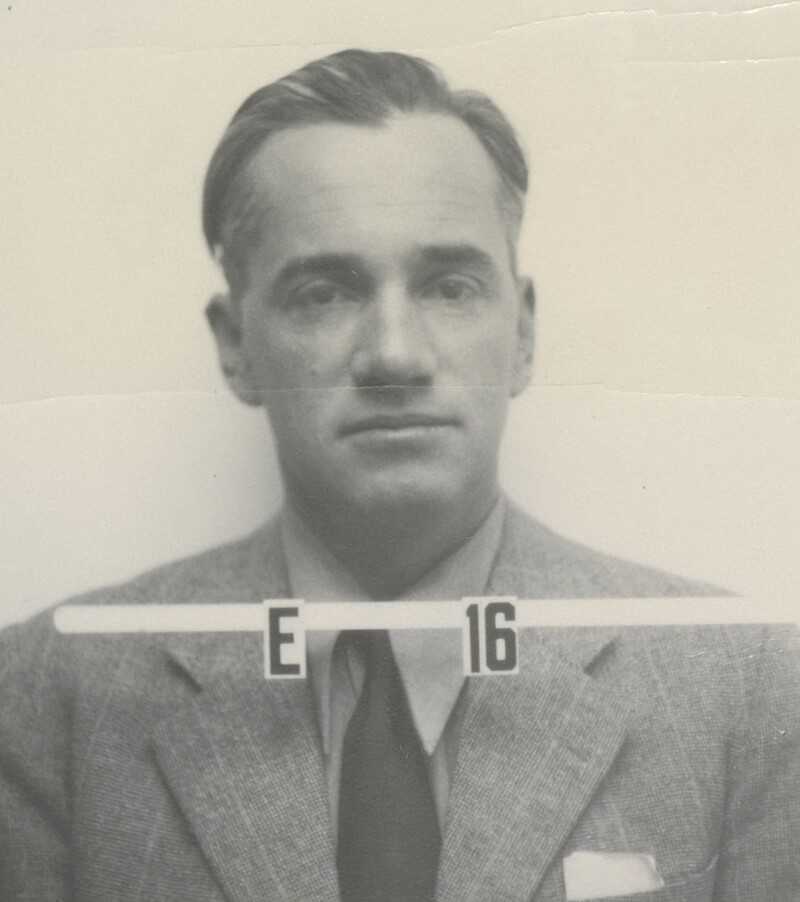  A black-and-white portrait of Otto Robert Frisch, a renowned nuclear physicist from the Manhattan Project era. He is depicted in formal attire with a serious expression. A clear badge, bearing the characters “E 16,” is visible on the image, possibly part of his official identification as a research team member.
