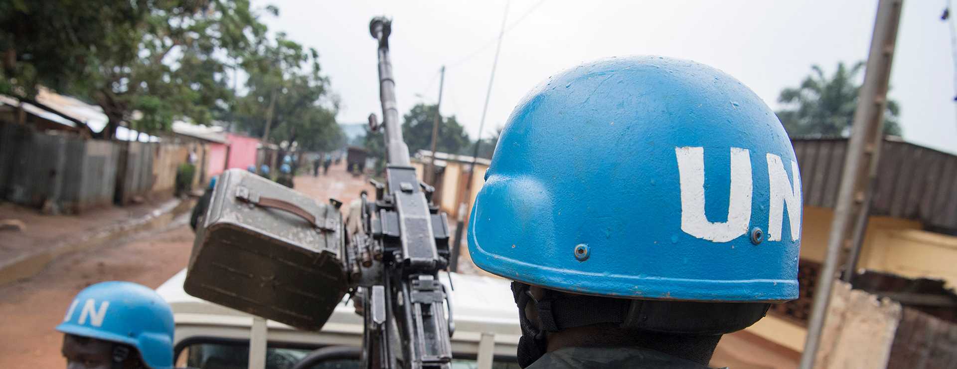 Photo taken outside on a sandy road. A person is pictured from behind, wearing a Blue Helmet with the letter UN on it holds a gun.